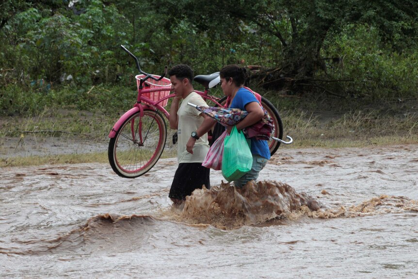 Two residents cross a swollen river carrying a bicycle and other belongings.