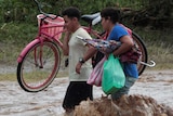 Two residents cross a swollen river carrying a bicycle and other belongings.