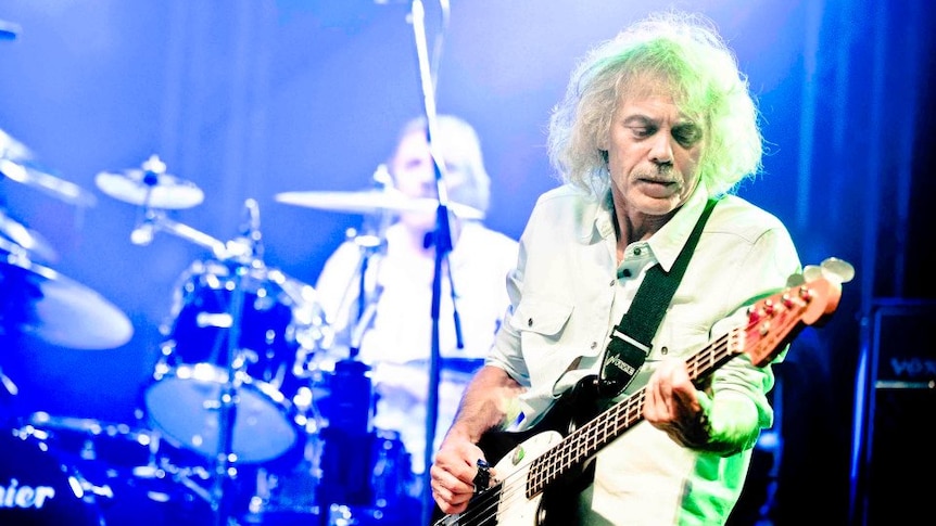 An elderly man with white curly hair plays bass guitar in front of a drummer on a brightly lit stage.