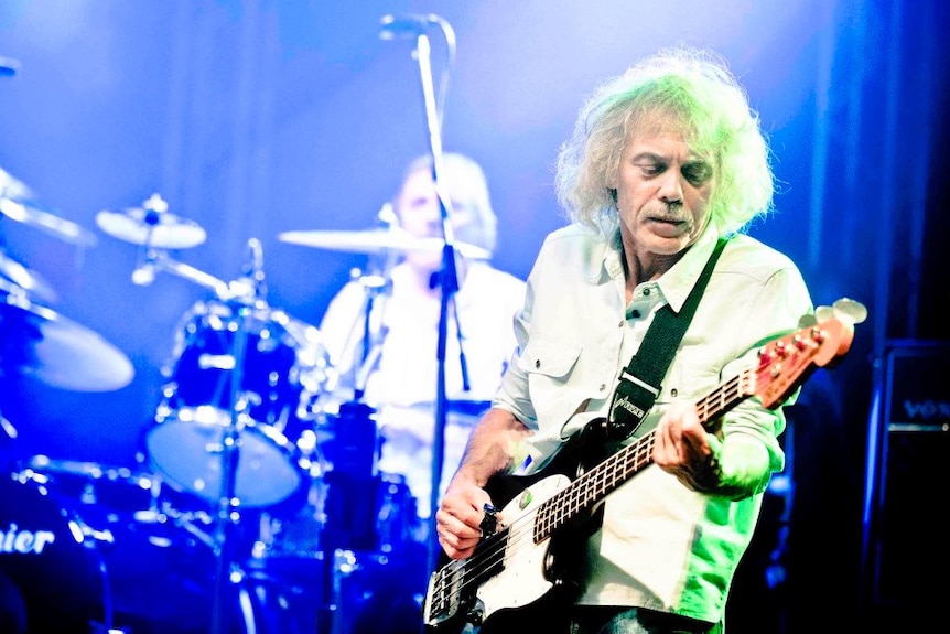 An elderly man with white curly hair plays bass guitar in front of a drummer on a brightly lit stage.