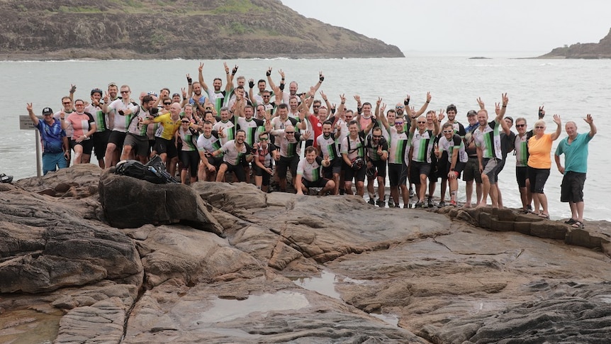 A group of men and women in lycra pants and t-shirts give the v sign as they stand on a rock platform next to water.