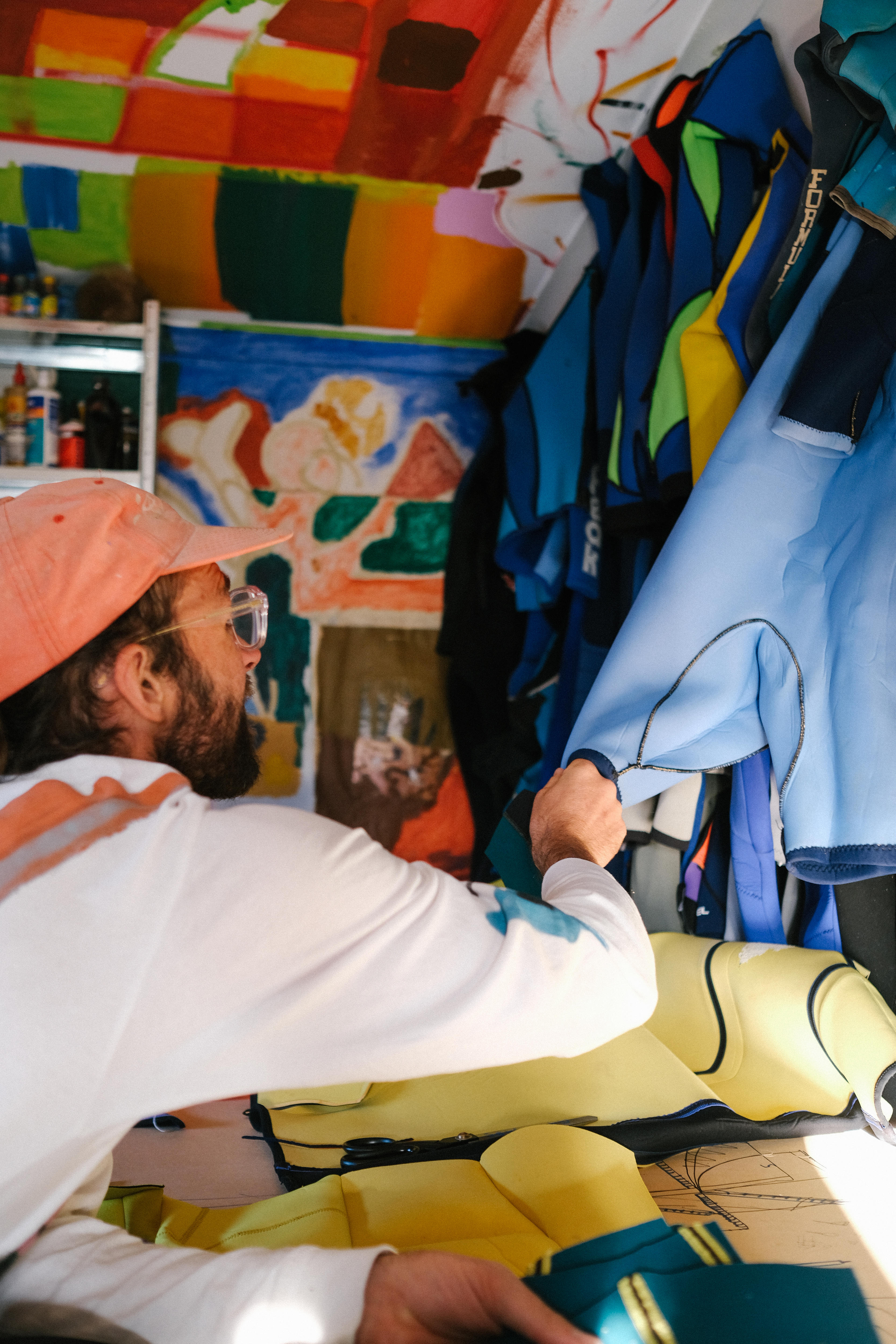 Artist wearing a red hat and white t-shirt leans across a table looking at a wetsuit on the wall