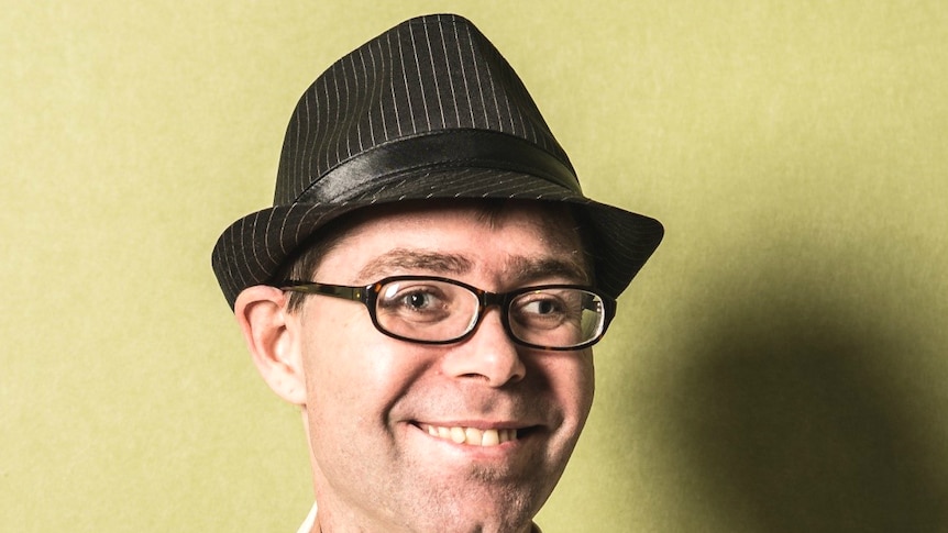 Head shot of man with glasses smiling and wearing a hat in front of a green background.