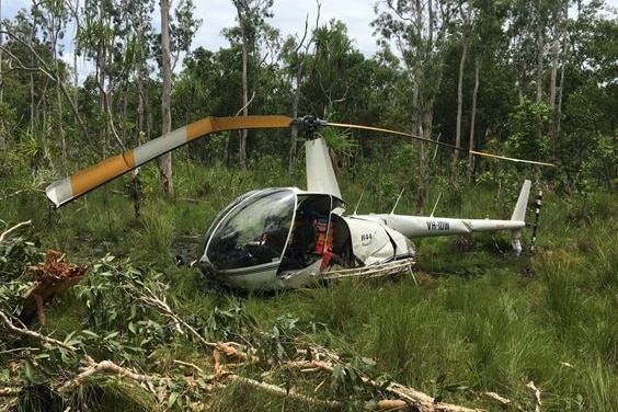 A small helicopter with damaged body and bend blades sit in bushland.