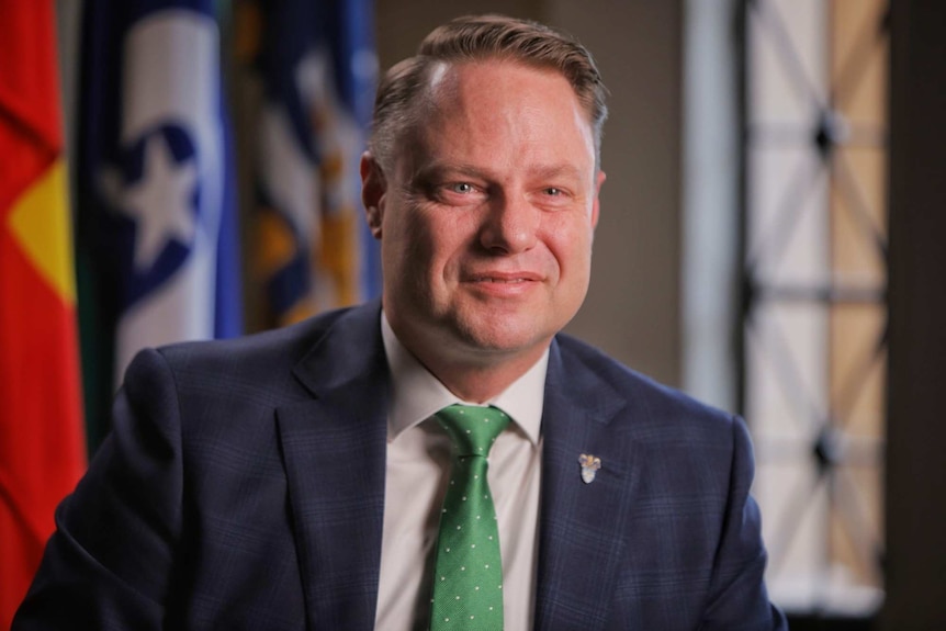 A man in a suit smiles with flags behind him.