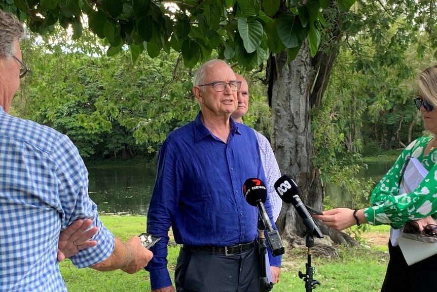 Professor Ross Garnaut stands in front of microphones in a leafy green area by some water, speaking.