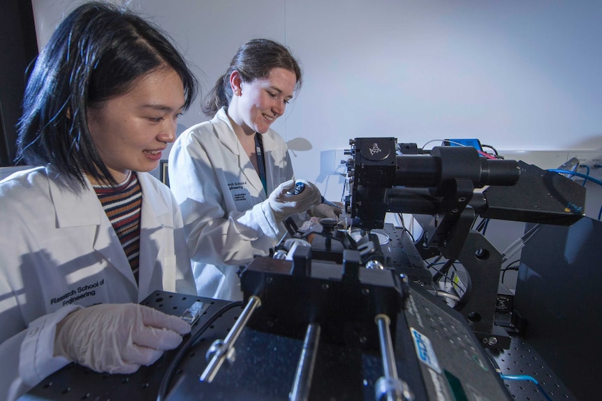 Two woman in lab coats operate a piece of diagnostic machinery.