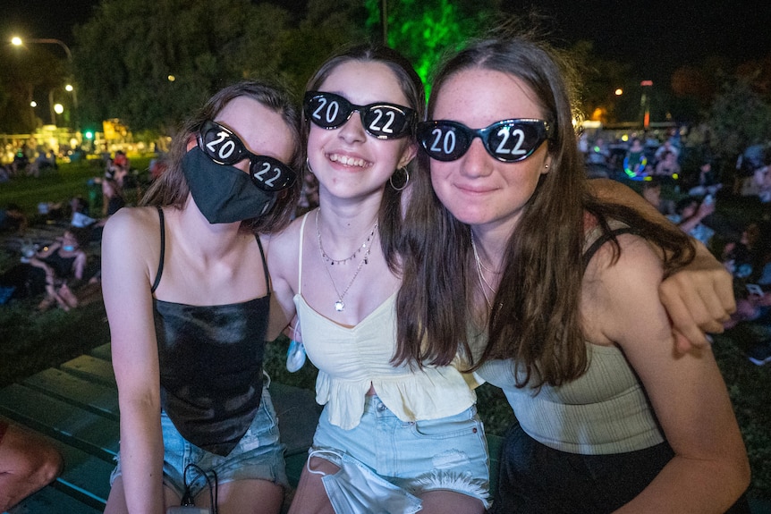 Three young girls sitting together wearing sunglasses in a park at night 