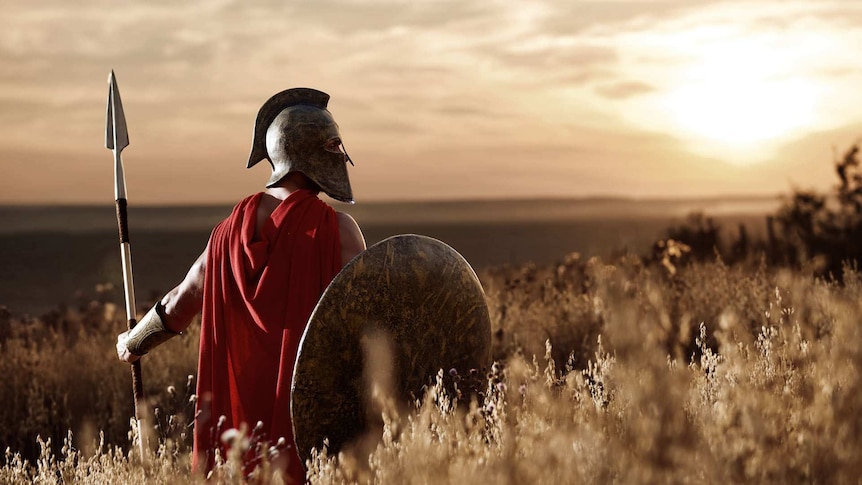 A muscular man, standing in a field, wearing a metal helmet and red cape, and holding a spear and a circular shield.