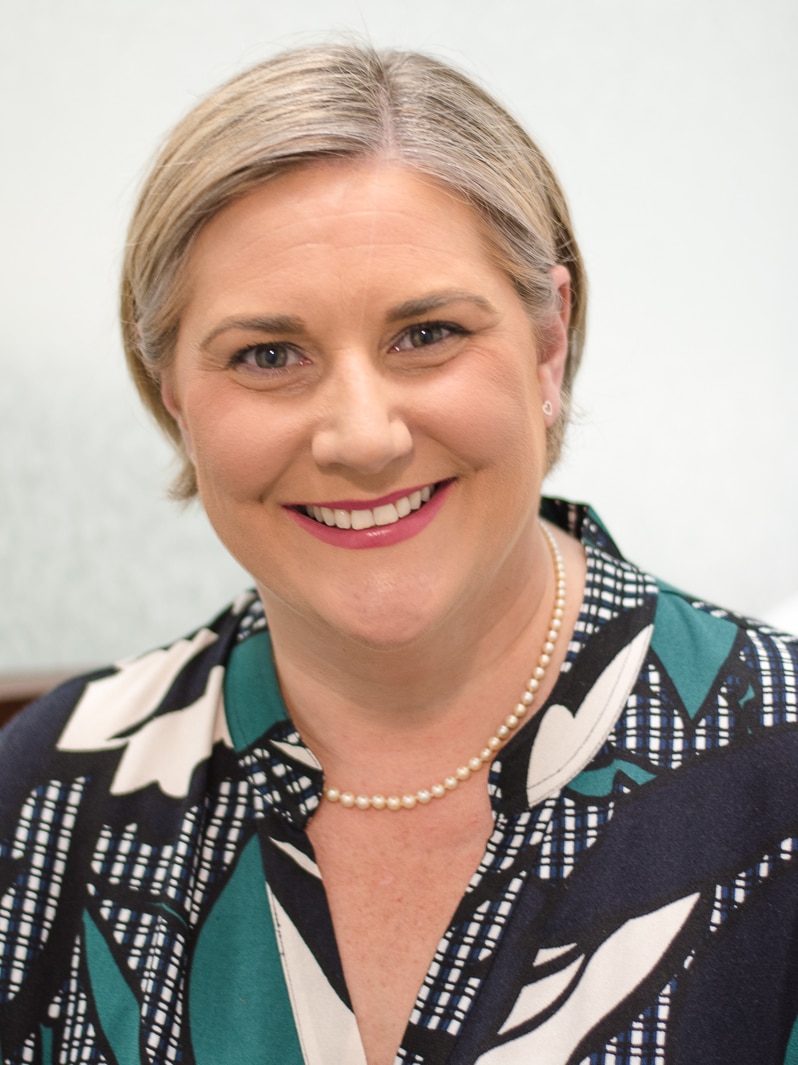 Hollie Hughes, photographed against a white background, smiles for a headshot. She is smiling and wearing a pearl necklace.