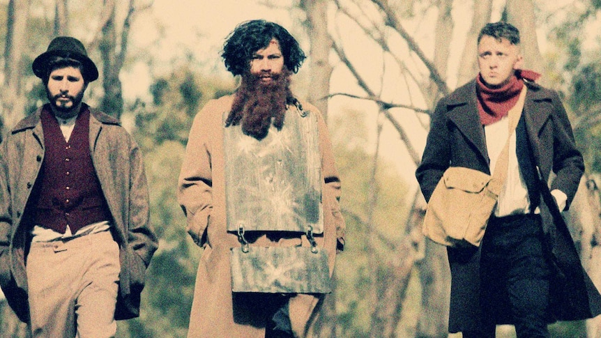 Three people dressed as the Ned Kelly Gang walk along a path through bushland toward the camera.