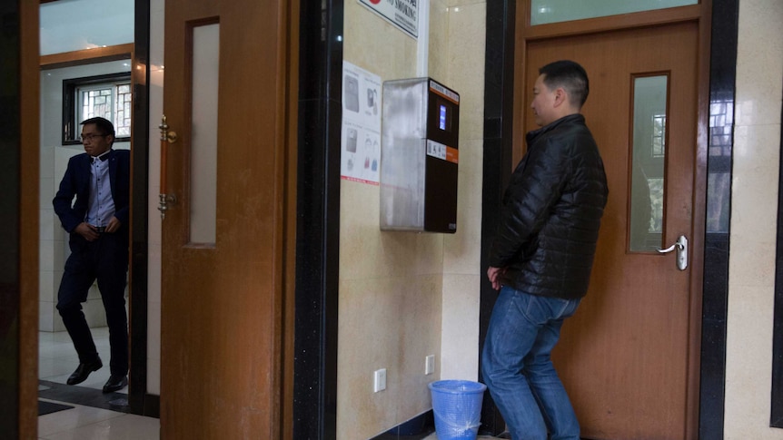 A man bends his knees to allow a toilet paper dispensing machine to scan his face at a toilet as another man leaves the toilet.