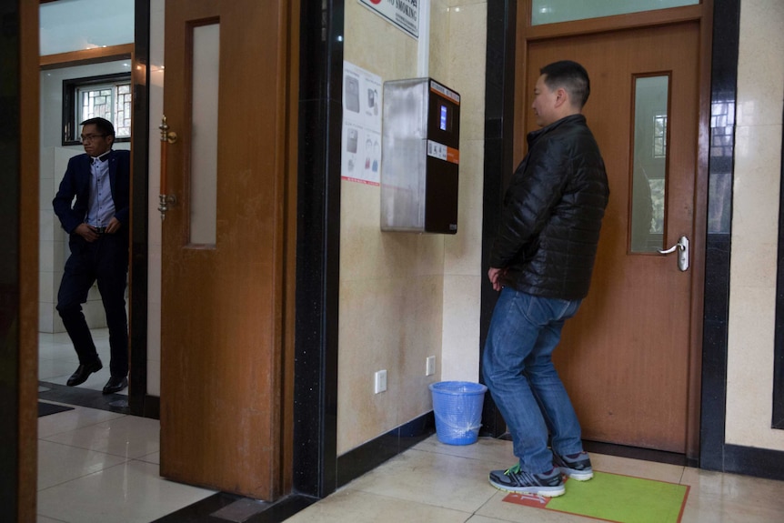 A man bends his knees to allow a toilet paper dispensing machine to scan his face at a toilet as another man leaves the toilet.