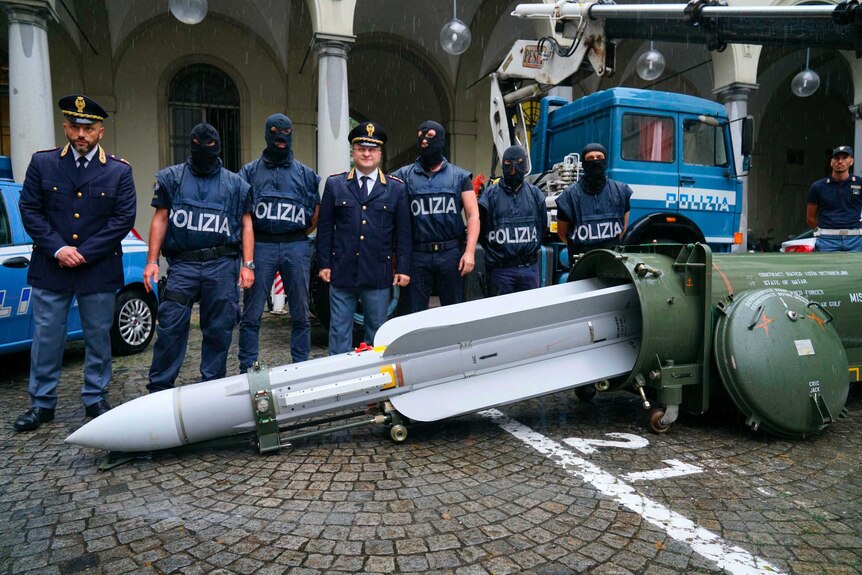 Police wearing balaclavas stand behind a large missile laying on the ground in front of them.