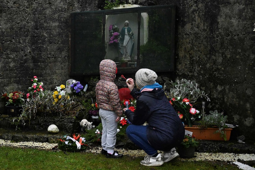 A woman and child stand and kneel as they look at a shrine.