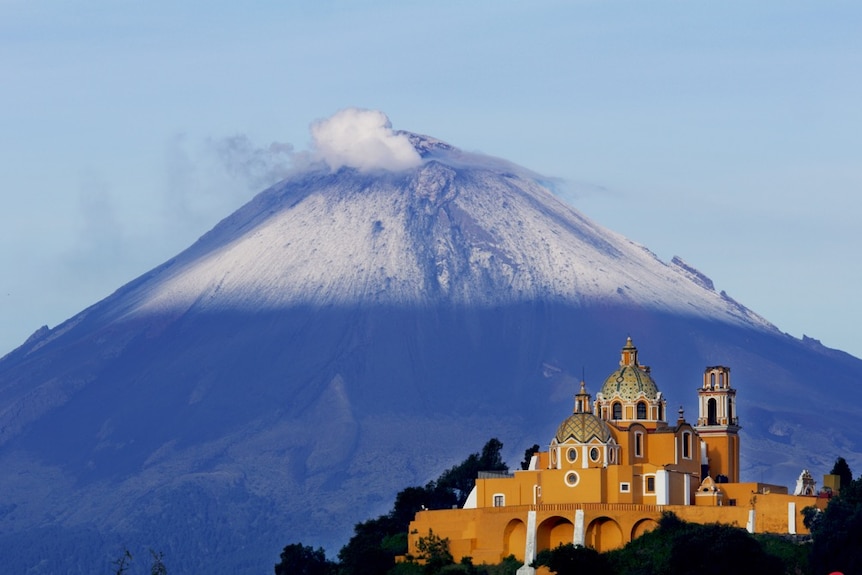 A snow-capped volcano with white smoke at its peak in the background with a mustard coloured castle in the foreground.