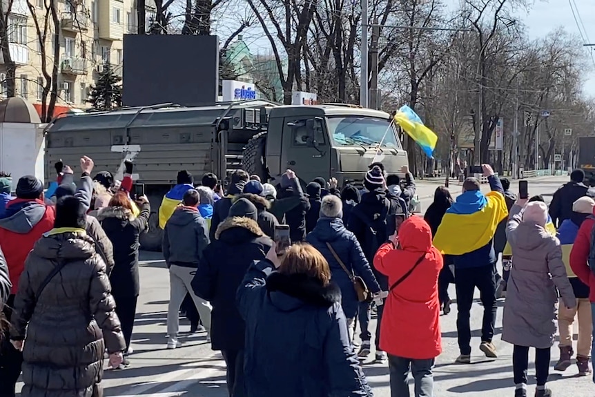 Protesters with Ukrainian flags stand in front of a military vehicle in the street. 