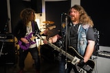 Two guitarists with long hair rehearse in a rock band, with one on the right wearing a denim jacket, singing into a microphone.