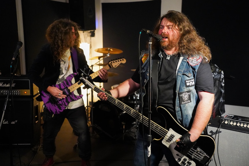 Two guitarists with long hair rehearse in a rock band, with one on the right wearing a denim jacket, singing into a microphone.