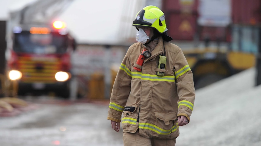 A firefighter wears a breathing mask, with fire trucks in the background.