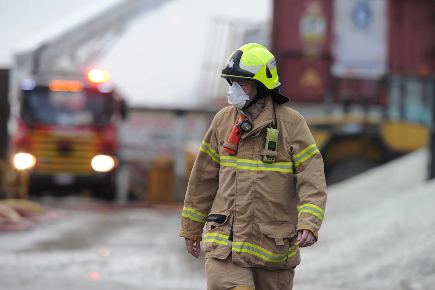 A firefighter wears a breathing mask, with fire trucks in the background.