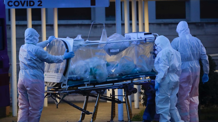 Health workers in hazmat suits wheel a person sealed inside a stretcher into a hospital