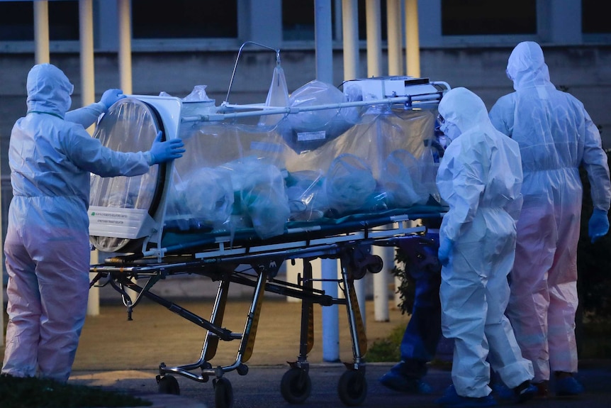 Health workers in hazmat suits wheel a person sealed inside a stretcher into a hospital