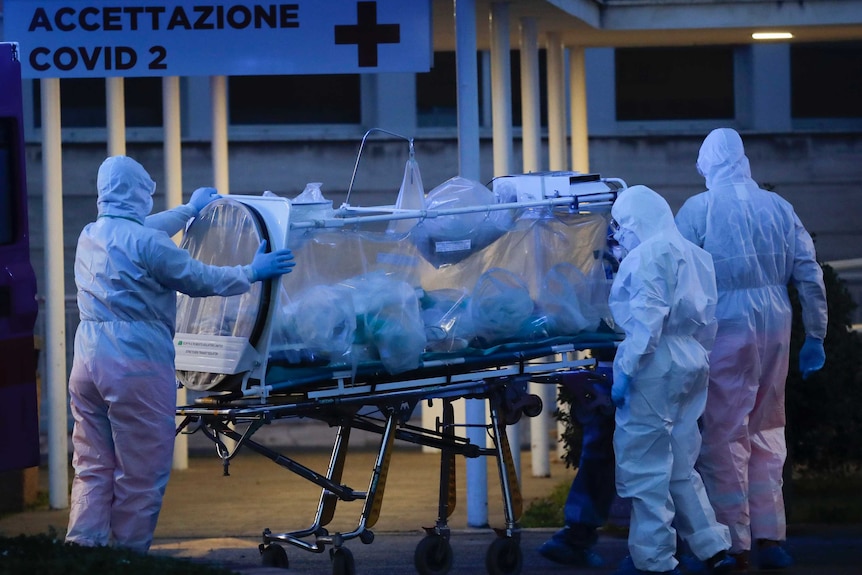 Health workers in hazmat suits wheel a person sealed inside a stretcher into a hospital