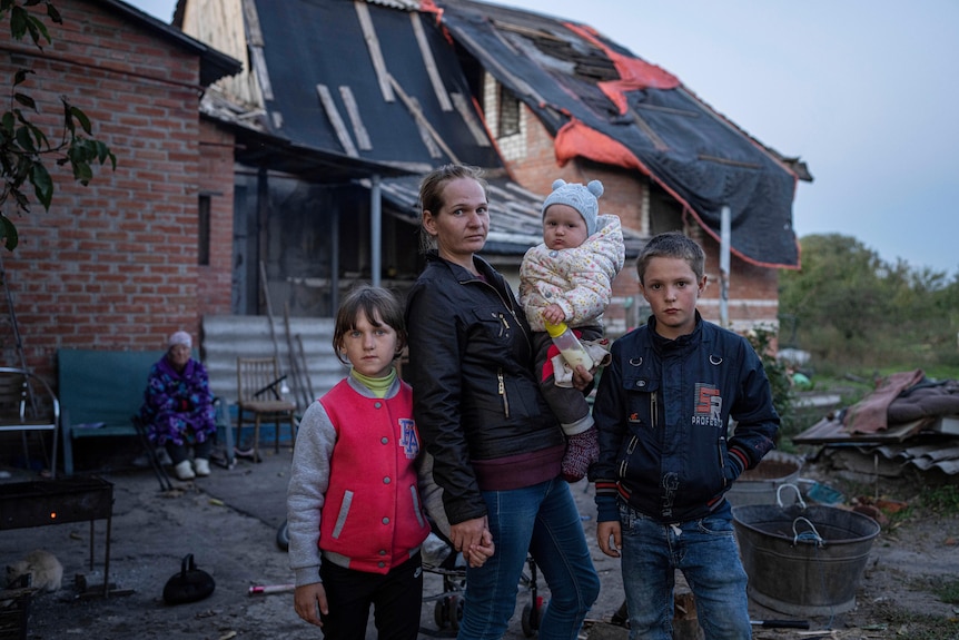 A woman with a baby stands with two older children on either side in the backyard of a destroyed house.