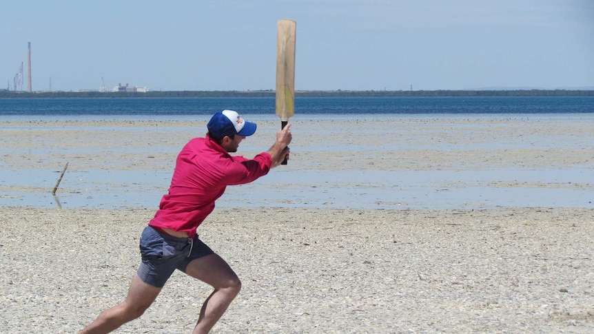 A man playing a cricket shot on the beach with the Port Pirie skyline in the background.