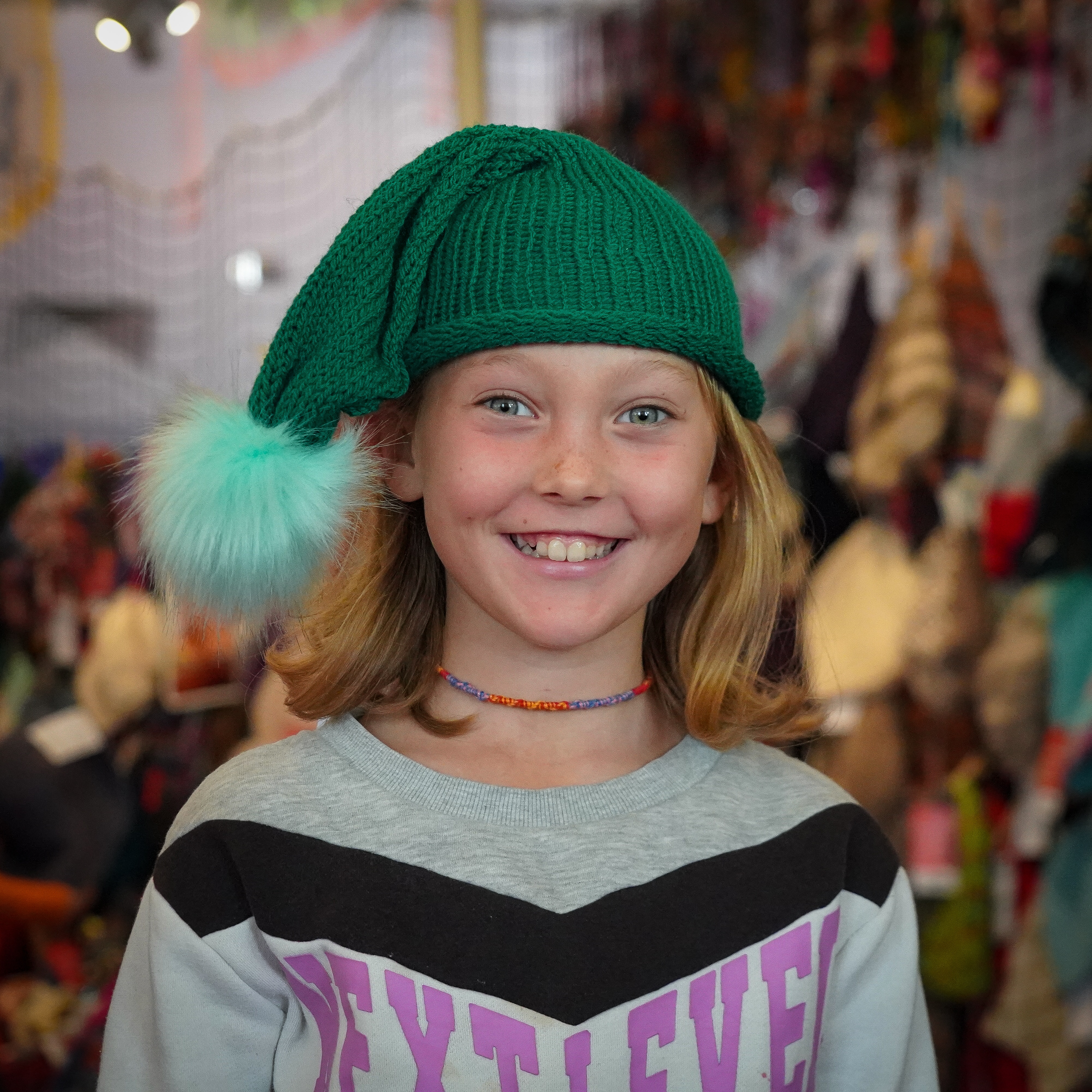 A young girl smiles at the camera while wearing a green knitted beanie with a fluffy pompom attached to it.