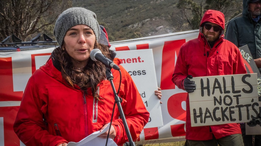 Adrien Butler stands in front of a microphone, wearing a grey beanie, red raincoat, with protest signs behind her.