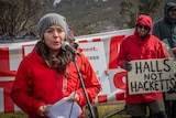 Adrien Butler stands in front of a microphone, wearing a grey beanie, red raincoat, with protest signs behind her.