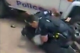 Two police officers hold down an Aboriginal man with a police vehicle in the background