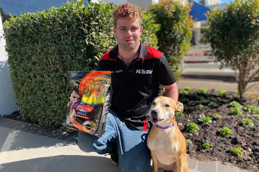 A man crouches down holding his small brown and white dog and a bag of dog food. 