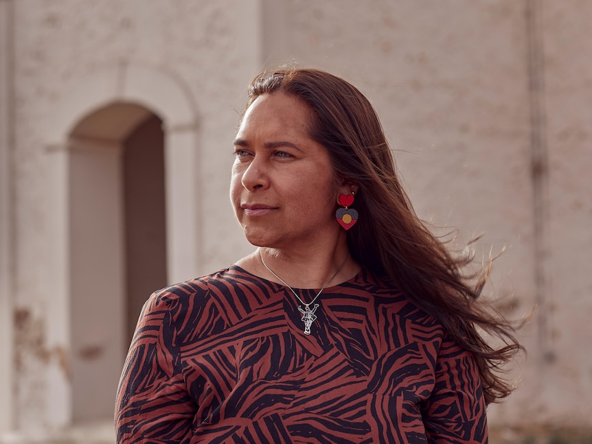 Tracey standing outside a sandstone building, wearing Indigenous flag earrings.