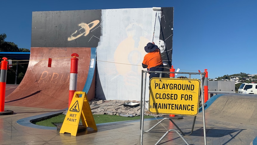 A council worker paints over a mural at a skate park with closed signs in the foreground.