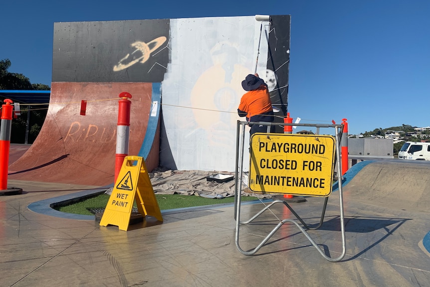 A council worker paints over a mural at a skate park with closed signs in the foreground.