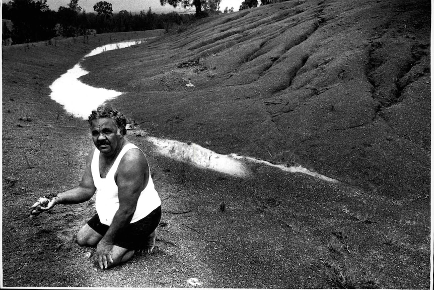 Image of a man kneeling on the ground with mounds of asbestos tailing waste around him. He is wearing a white singlet.