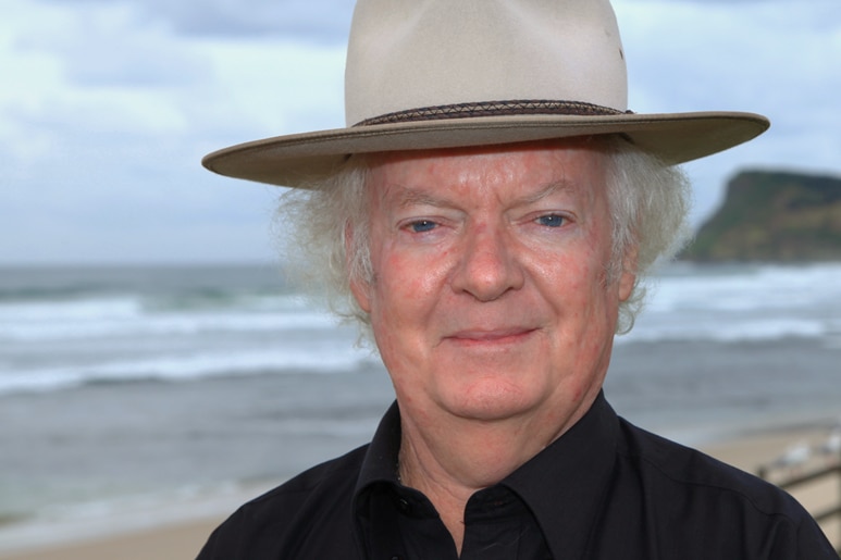 A portrait shot of a man in a black shirt and felt hat with the beach in the background