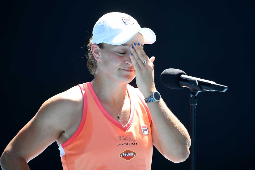 Ash Barty closes her eyes and touches her forehead while speaking into a microphone on a tennis court.
