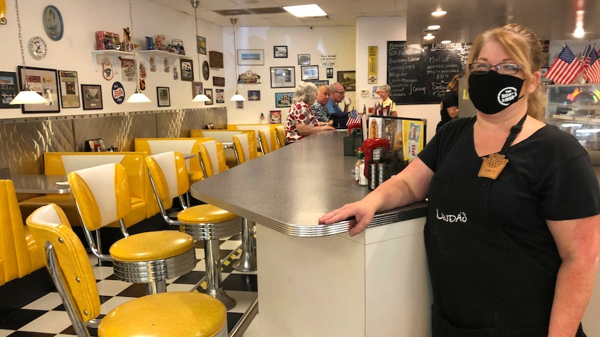 A woman in a face mask stands at the counter of a nearly empty diner with US flags on the wall.