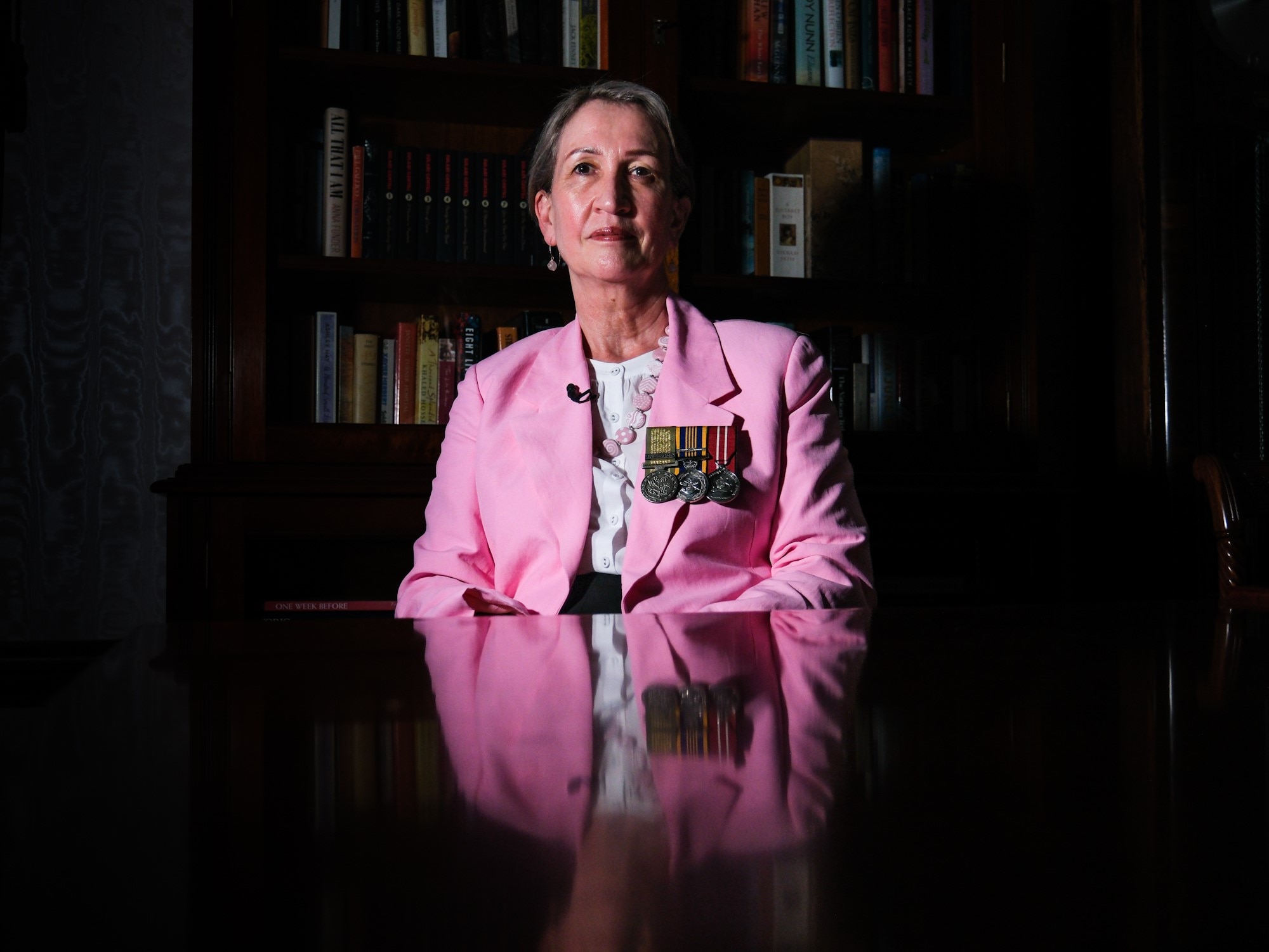 a woman wearing war medals on the lapel of her jacket sitting down behind a table and looking at the camera