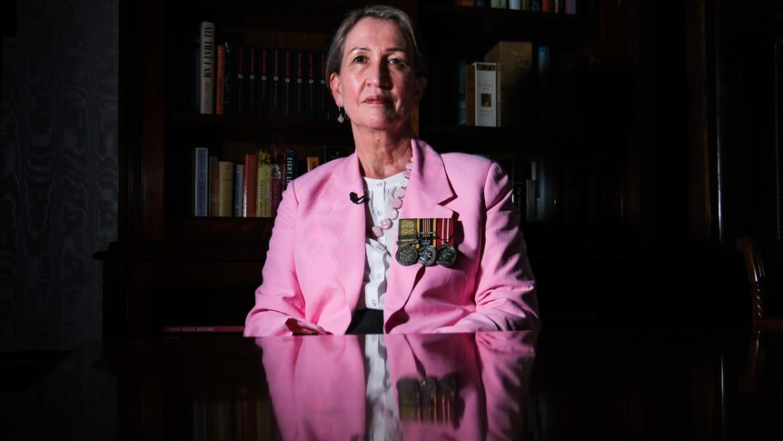 a woman wearing war medals on the lapel of her jacket sitting down behind a table and looking at the camera