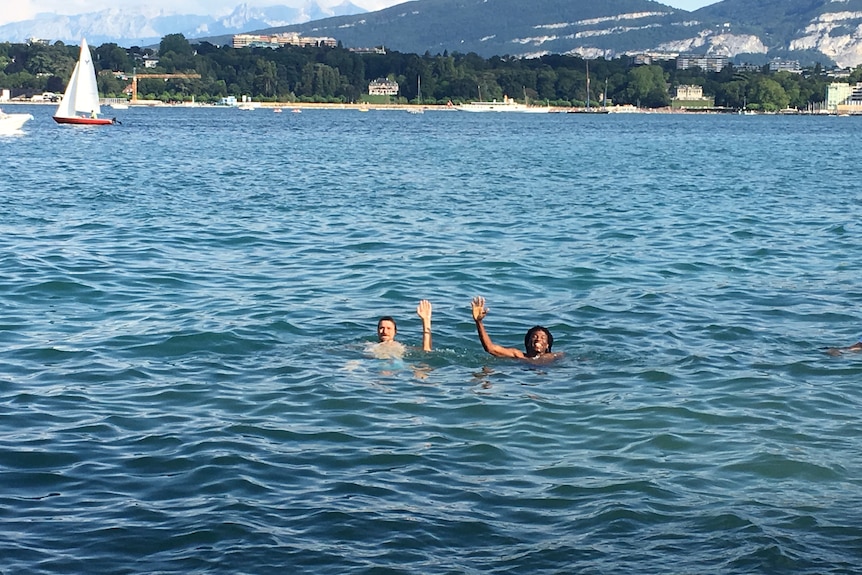 Two men wave while swimming in lake.