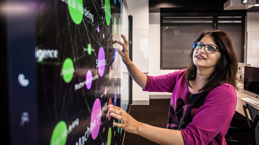 An image of Professor Shazia Sadiq standing facing a wall with colourful circles and data