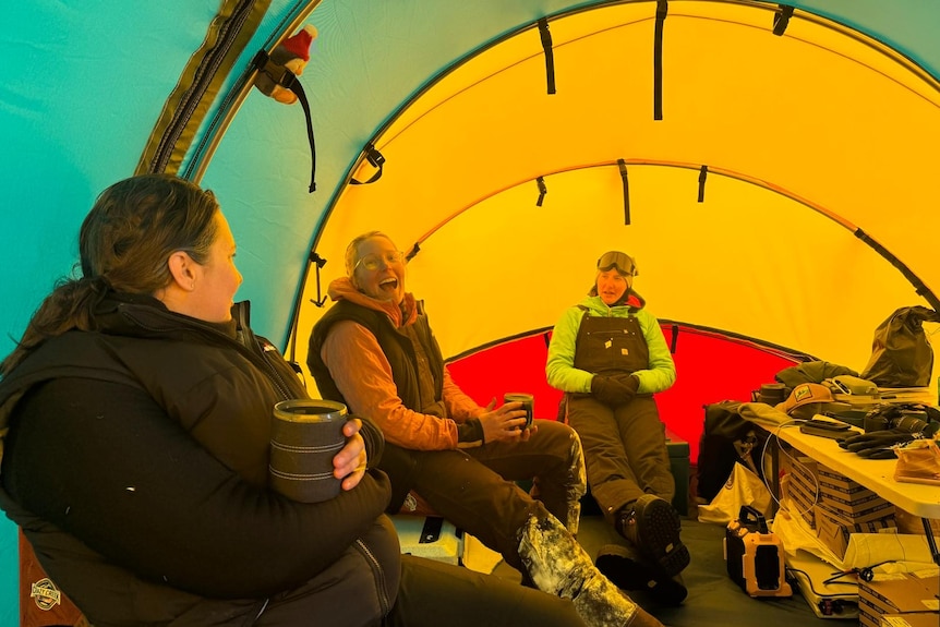 Three women sit inside a yellow and green tent. They're wearing thick winter gear and talking amongst themselves.