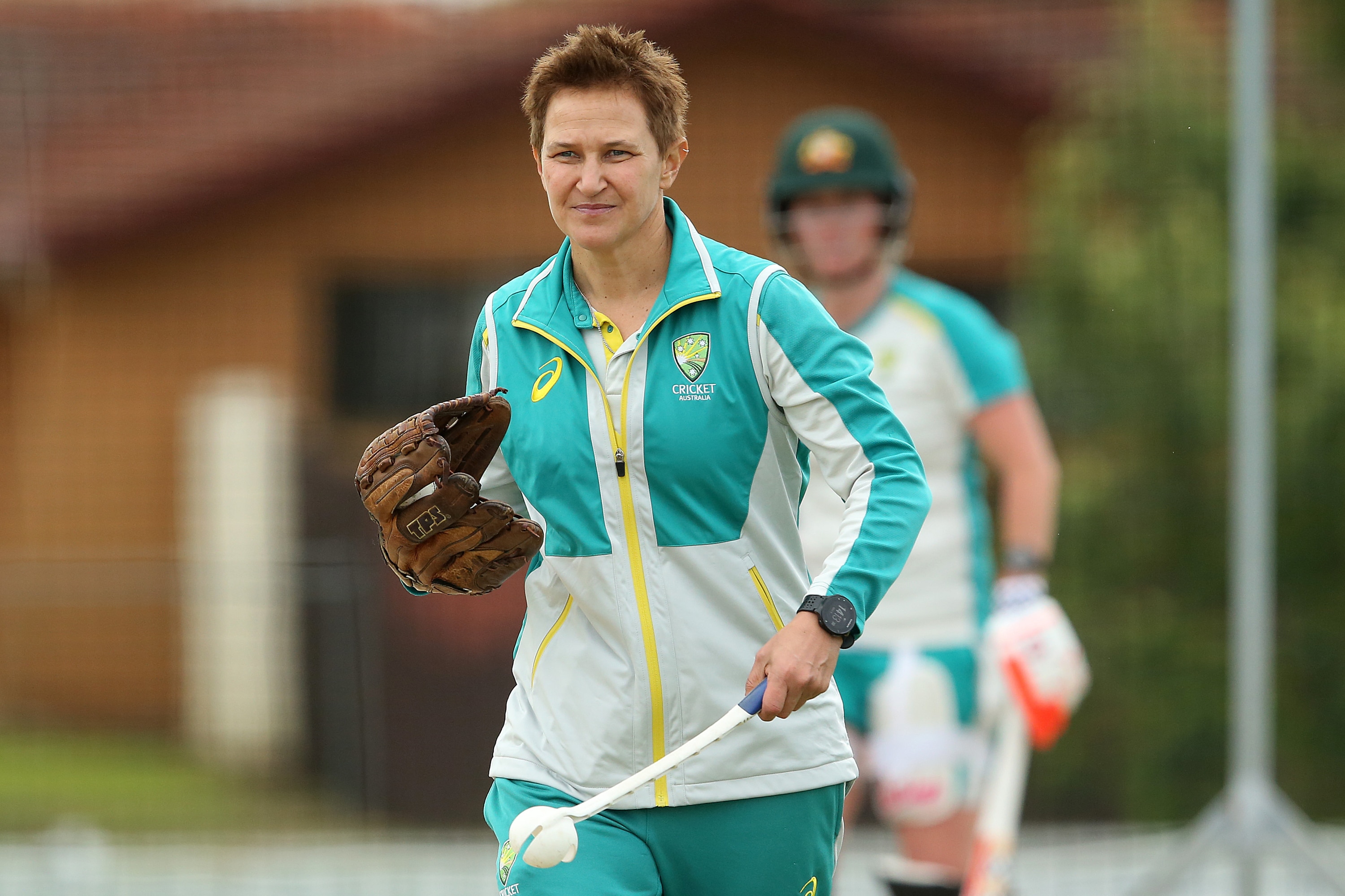 A coach walks across a ground, with a baseball glove on one of her hands, and a ball in a claw for throwdowns on the other.
