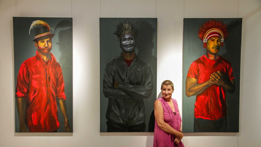 Joan Winter stands with three large portrait paintings from the Fish Hooks and Moving Trees exhibition at Bundaberg Regional art