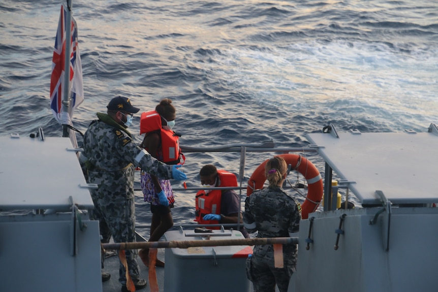 Defence personnel wearing masks help people in life vests aboard a ship.